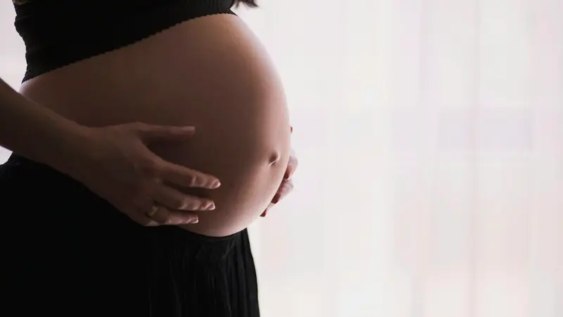 Expectant mother in soft natural window light, resting her hands protectively on her pregnant belly in a moment of quiet reflection.