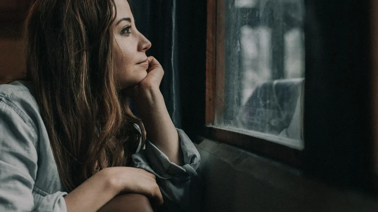 A woman sits quietly by a window, chin resting on her hand, gazing out in soft muted light in a moment of thoughtful reflection.