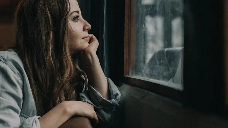 A woman sits quietly by a window, chin resting on her hand, gazing out in soft muted light in a moment of thoughtful reflection.