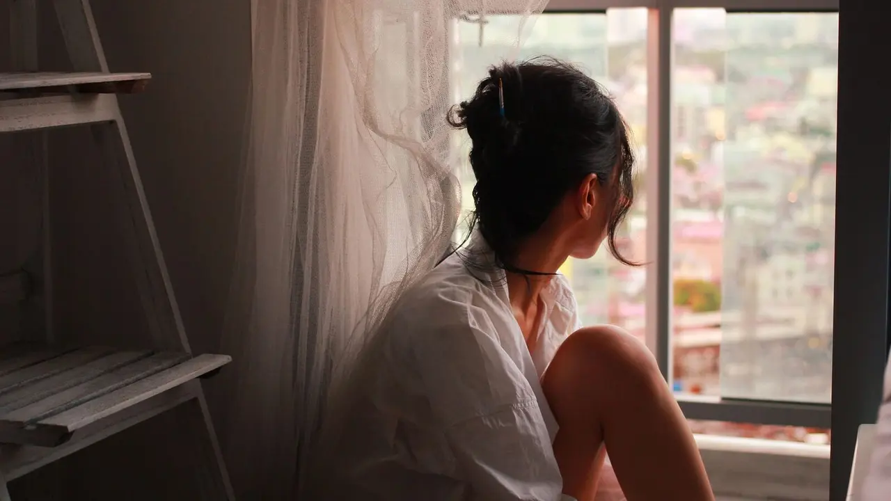 Woman sitting quietly by a window in soft natural light, looking thoughtful in a calm, reflective mood.